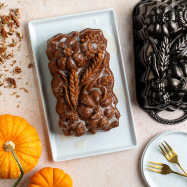 Overhead image of glazed Sunflower Pumpkin Loaf cake on white plate surrounded pan, pumpkins and crushed pecans