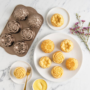 Six Plain Floral Cakelets on white plate, two Floral cakelets on smaller white plates, flowers and pan displayed next to plates