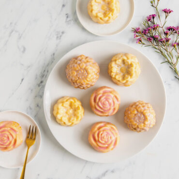 Six frosted Floral Cakelets on white plate, two Floral cakelets on smaller white plates with flowers displayed next to plates
