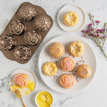 Six frosted Floral Cakelets on white plate, two Floral cakelets on smaller white plates, flowers and pan displayed next to plates