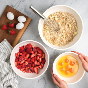 Nordic Ware 3 piece white prep and serve 5 quart, 3.5 quart and 2 quart bowls on kitchen countertop filled with strawberries, eggs and dough