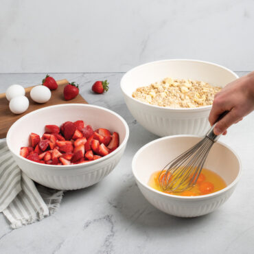 Nordic Ware 3 piece white prep and serve 5 quart, 3.5 quart and 2 quart bowls on kitchen countertop filled with strawberries, eggs and dough