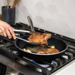An image of a black 12-in skillet with a white speckled pattern being used on the stovetop, cooking porkchops.