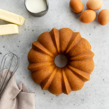 Golden bundt cake on countertop with baking ingredients including butter, eggs, vanilla cake mix, and whisk
