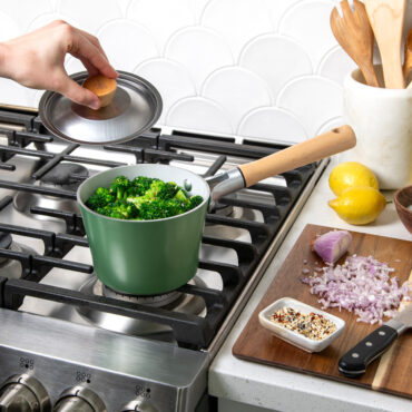 Cardamom 1.5Qt Saucepan with broccoli in pan on stovetop, hand holding lid over pan