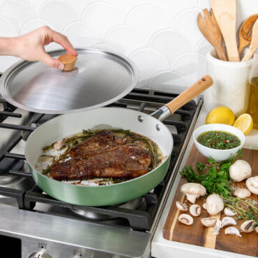 Nordic Ware Cardamom 12" nonstick frying pan with lid, cooking seared steak on stovetop with herbs nearby