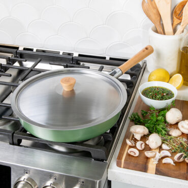 Nordic Ware Cardamom 12-inch nonstick frying pan with glass lid on stovetop, surrounded by cooking ingredients