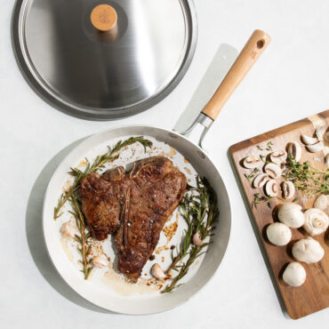 Nordic Ware Cardamom 12-inch nonstick frying pan with lid, containing seared steak, ingredients on cutting board