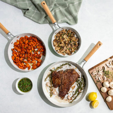Overhead mage of Cardamom 3 piece fry pans with steak, carrots and mushrooms in pans, on countertop with various ingredients