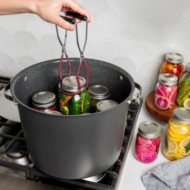 20 Qt Stock Pot with lid on stovetop with jars of veggies inside stock pot; hand using tongs to pull one jar out