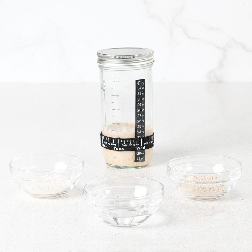 Image of starter jar with sourdough starter in it with bowls of flour water and salt on surface