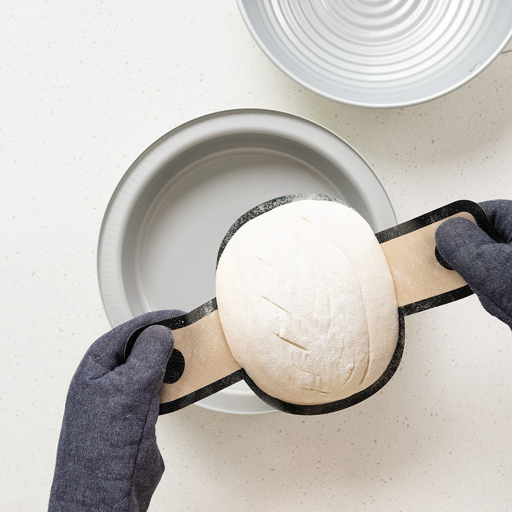 Overhead image of hand lifting scoured sourdough bread using lifting mat and placing in pan