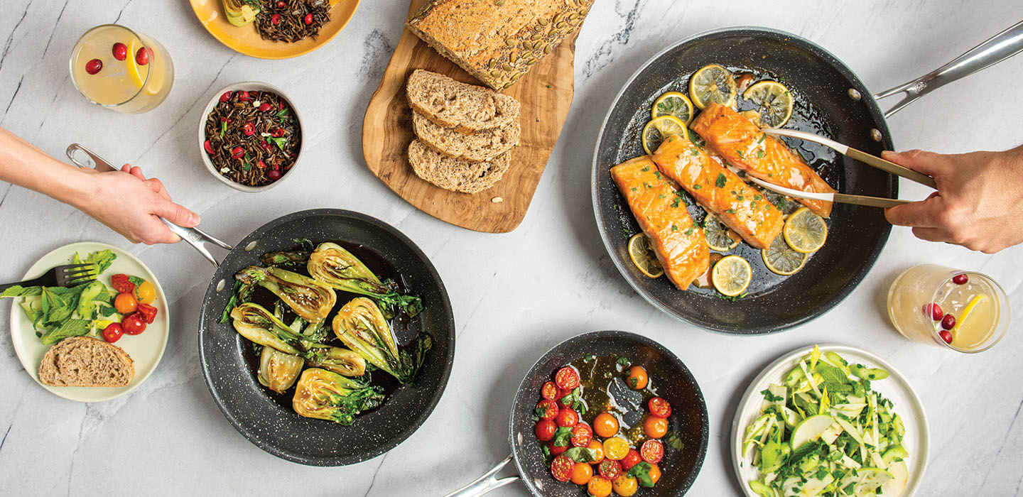 Dinner scene of salmon and vegetables being serves on a table in Basalt Nonstick Ceramic Cookware. Two hands are coming in to serve the food onto plates. 