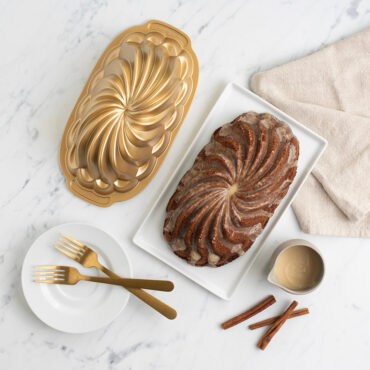 Pirouette Loaf cake glazed on plate next to pan, cup of frosting, and cinnamon sticks, plate with two forks