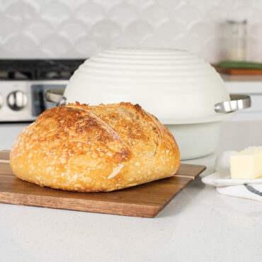 Freshly baked sourdough bread sitting on cutting board atop a kitchen counter, stick of butter next to it with pan and lid behind