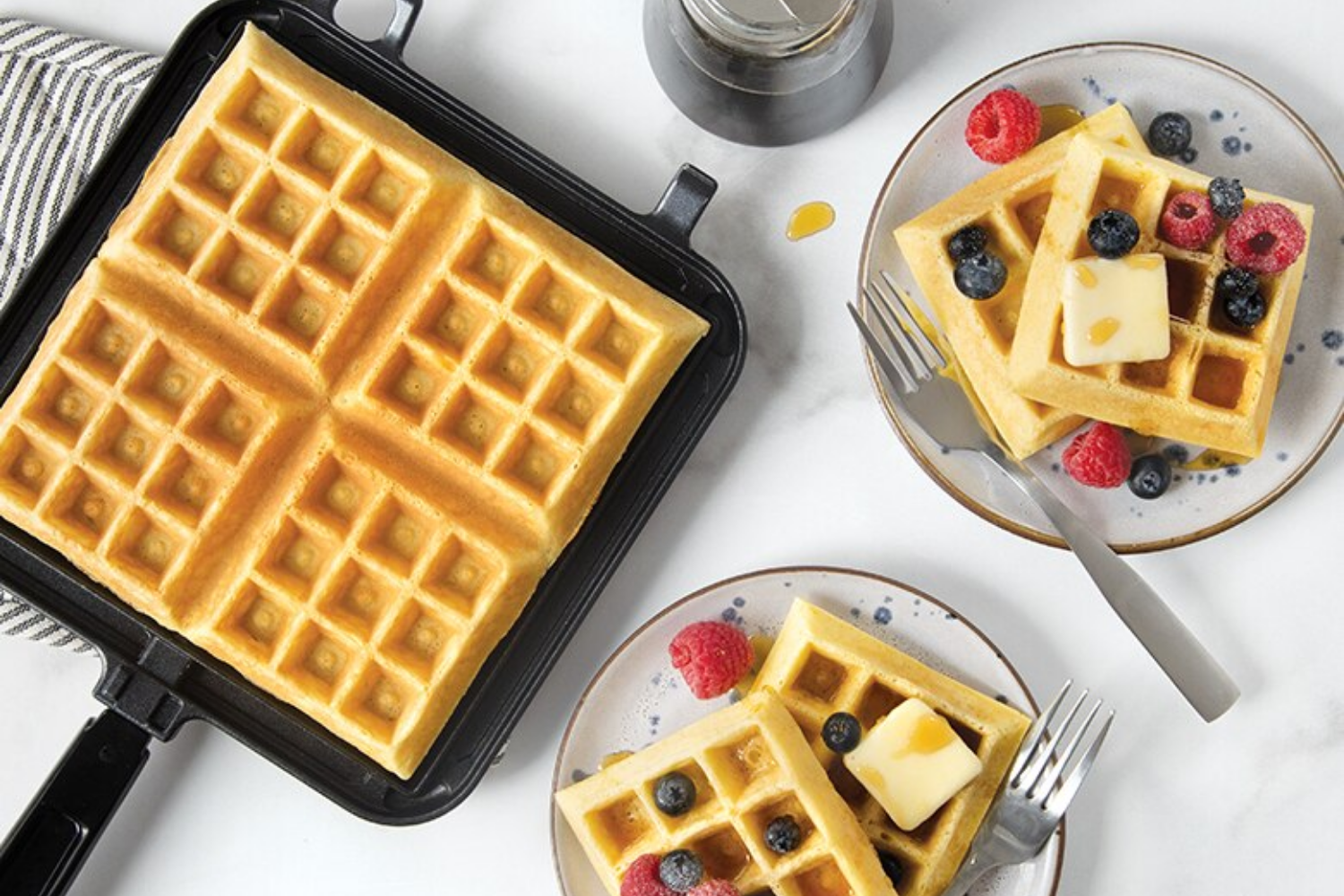 Original Stovetop Belgian Waffler with a cooked waffle inside of it and two plates of waffles with berries on top next to it.