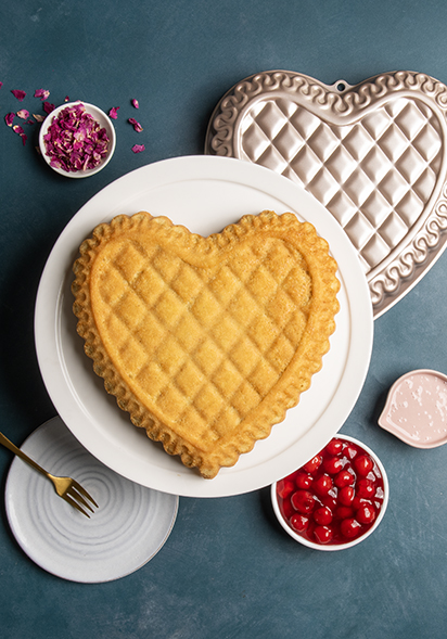 Image of baked quilted heart cake on a cake plate with pan and ingredients around it.