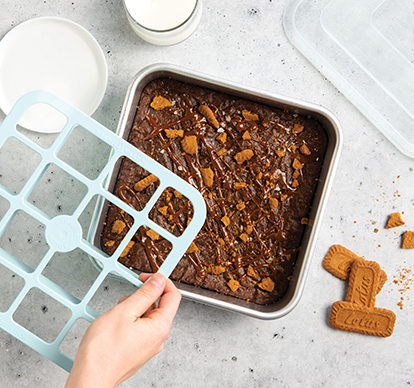 image og baked brownies in 9 inch brownie pan with hand about to cut with brownie cutter.
