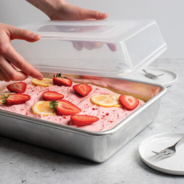 frosted cake with strawberry and orange slices in rectangular baking pan, hands holding lid over pan