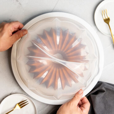 Clear plastic bundt cake keeper with domed lid containing golden baked brilliant bundt cake, marble background, overhead, hands closing lid