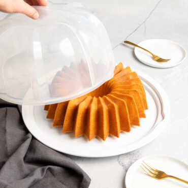 Clear plastic bundt cake keeper with domed lid containing golden baked brilliant bundt cake, hand holding lid over base