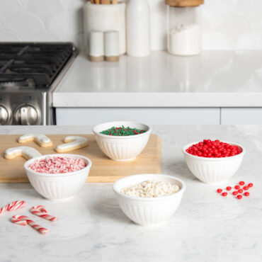 Four white mini bowls arranged on white countertop with crushed candy cane, red and green sprinkles, and red candies