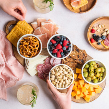 Four mini white bowls on a charcuterie board filled with snacks and cheese, two hands grabbing food in scene