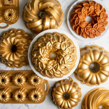 Laurel Wreath Bundt on white cake stand surrounded by various gold Bundt Pans and Laurel Wreath Bundt Cake