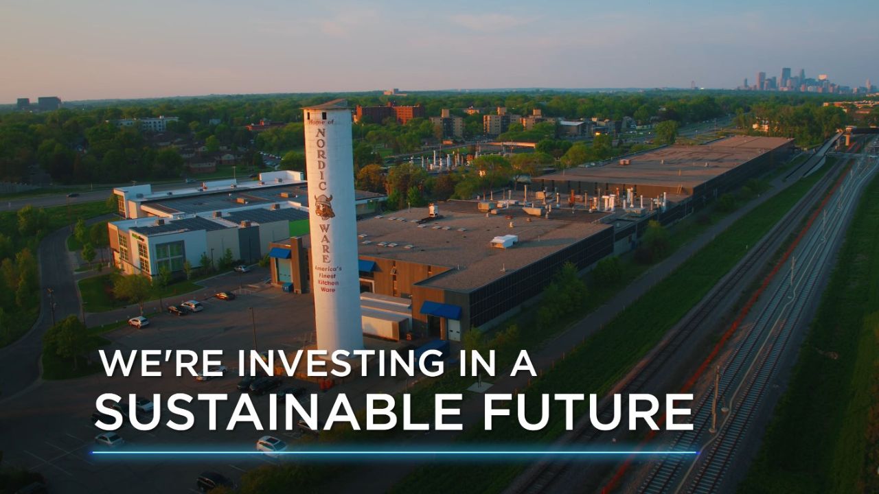 Image of Nordic Ware factory and solar panels at sunset with copy "We're investing in a sustainable future." Click to watch solar panel video
