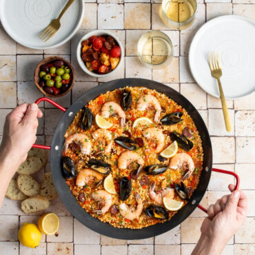 paella pan with saffron rice, shrimp, mussels, and vegetables, hands placing pan on tile countertop