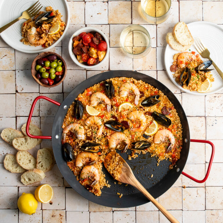  Image of spanish paella in 15" Paella Pan with Red Handles with a serving spoon and dinner plates in summer scene