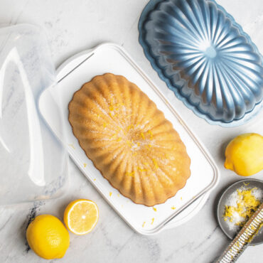 overhead image of lemon fluted loaf cake in keeper on marble countertop with lid off, lemons and pan scattered around