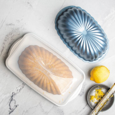 overhead image of lemon fluted loaf cake in keeper on marble countertop, lid closed, lemon and pan placed nearby