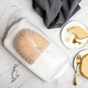 overhead image of lemon fluted loaf cake cut in half in keeper on marble countertop, lid closed, with small plates with cake slices