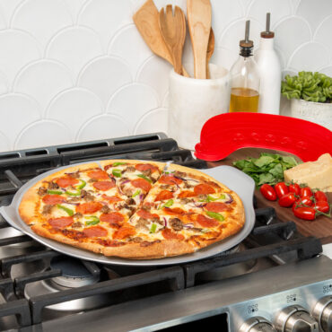 lifestyle scene of a cut supreme topping pizza on pizza pan placed on stovetop with rock the boat pizza cutter staged on cutting board with ingredients