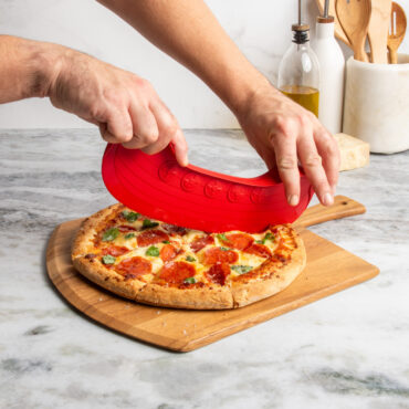 lifestyle scene of hands using rock the boat pizza cutter to cut a supreme topping pizza on cutting board placed on marble countertop