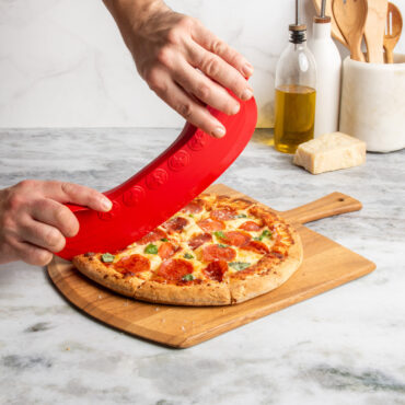 lifestyle scene of hands using rock the boat pizza cutter to cut a supreme topping pizza on cutting board placed on marble countertop