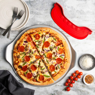 overhead lifestyle scene of sliced supreme topping pizza on pizza pan staged on marble countertop next to rock the boat pizza cutter and various seasonings