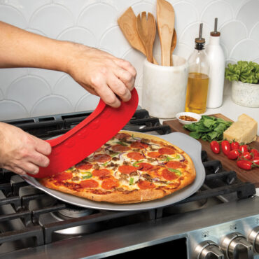 lifestyle scene of hands using rock the boat pizza cutter to cut a supreme topping pizza on cutting board placed pizza pan placed on stovetop