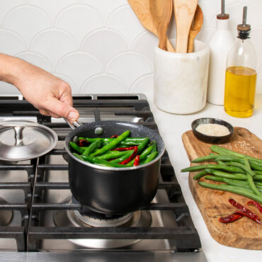 hand grabbing handle of basalt 1.5qt saucepan filled with string beans on stovetop, lid placed behind pan and cutting board with ingredients next to stove on countertop