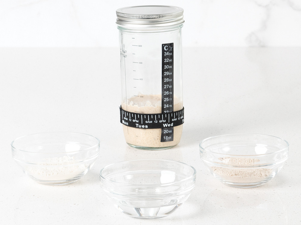 Sourdough Starter jar filled with starter with components to feed starts in glass bowls surrounding it.
