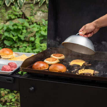 hand holding cheese melting dome over outdoor grill with burger patties and buns