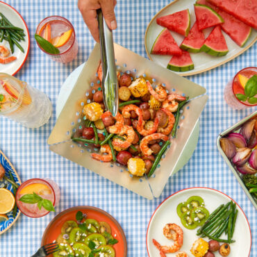 nordic ware grill basket staged on table with plates of fruits and veggies, hand with tongs grabbing a piece of shrimp out of basket