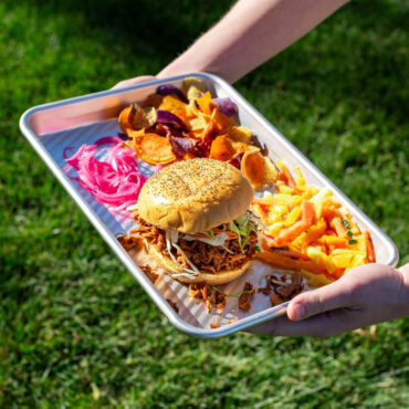 pulled pork sandwich, fries, and chips being carried in an outdoor setting on nordic ware aluminum grill tray