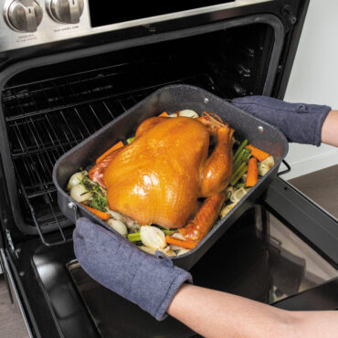 hands wearing oven mitts pulling Nordic Ware Verde Roaster with freshly baked turkey and veggies out of oven