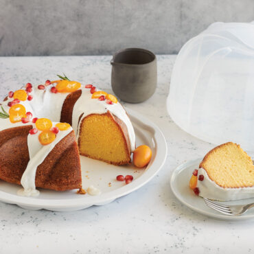Red Nordic Ware aluminum bundt pan with clear plastic cake keeper dome, frosted bundt cake inside